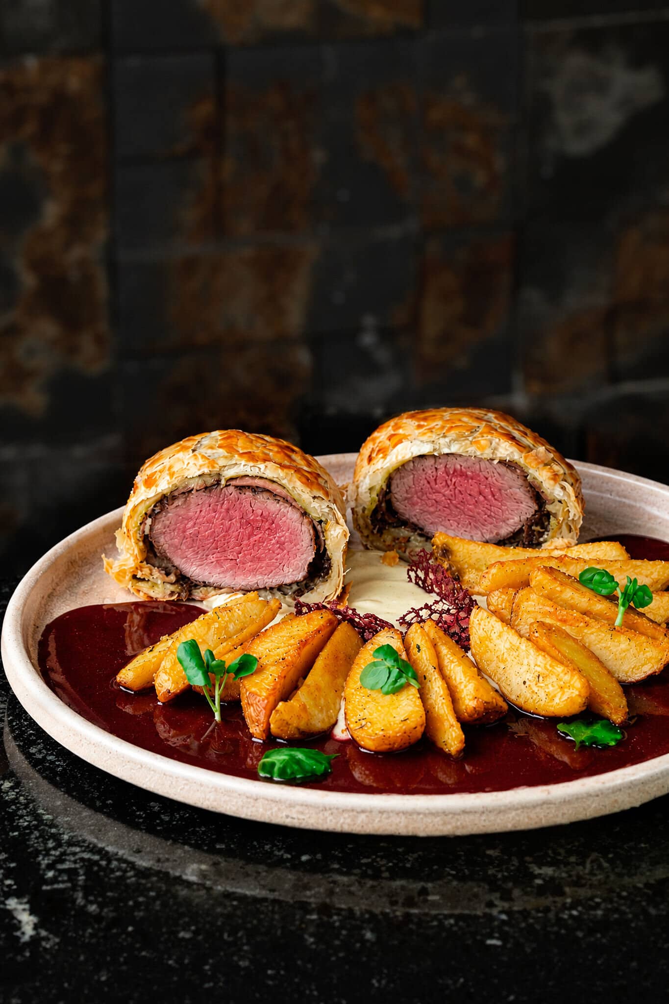 Beef Wellington cortado ao meio, filé rosado dentro da massa folhada, com batatas rústicas e molho escuro.