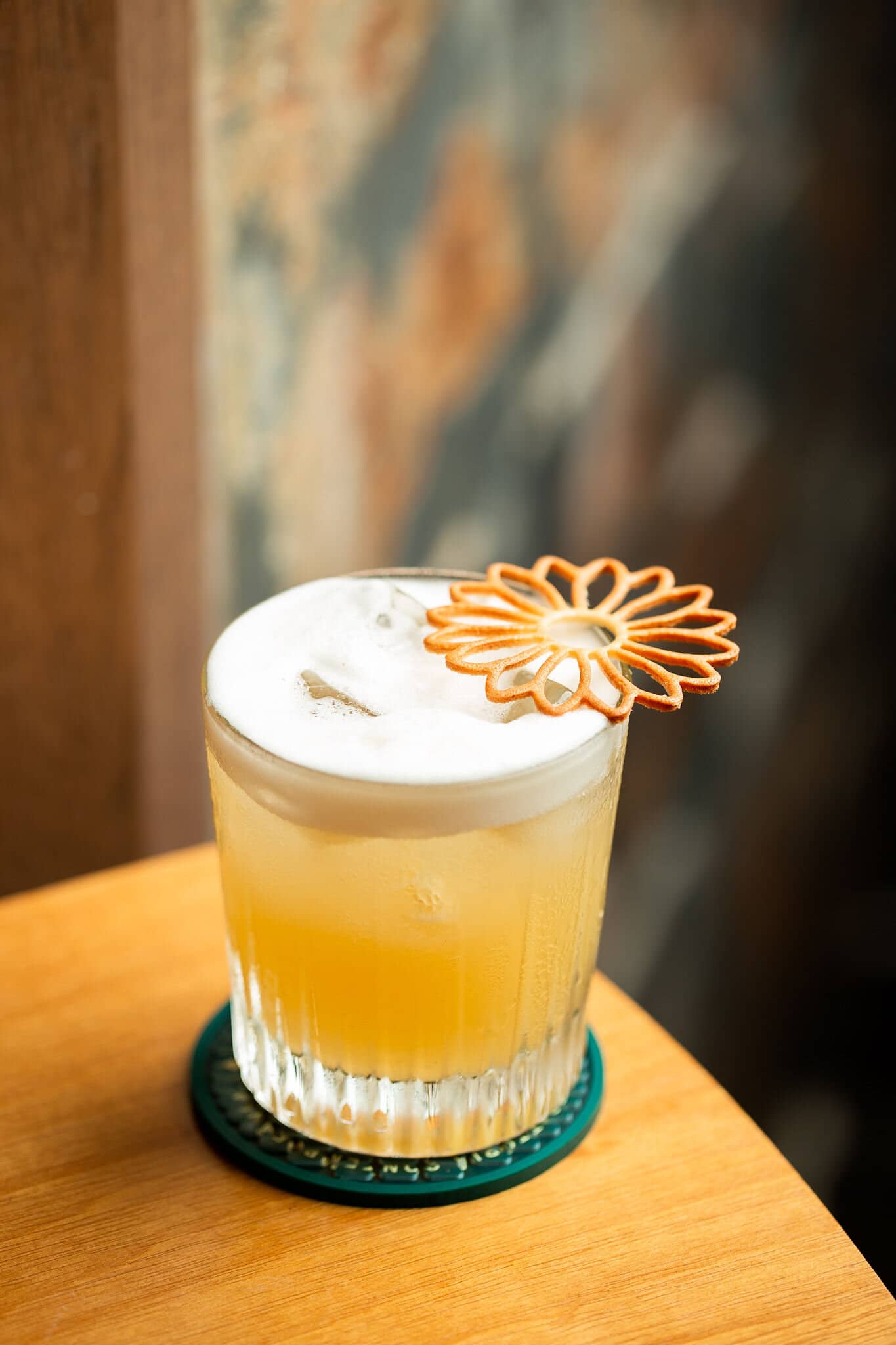 Drinque de espuma clara em copo baixo, com uma flor de açúcar caramelizado, sobre mesa de madeira.