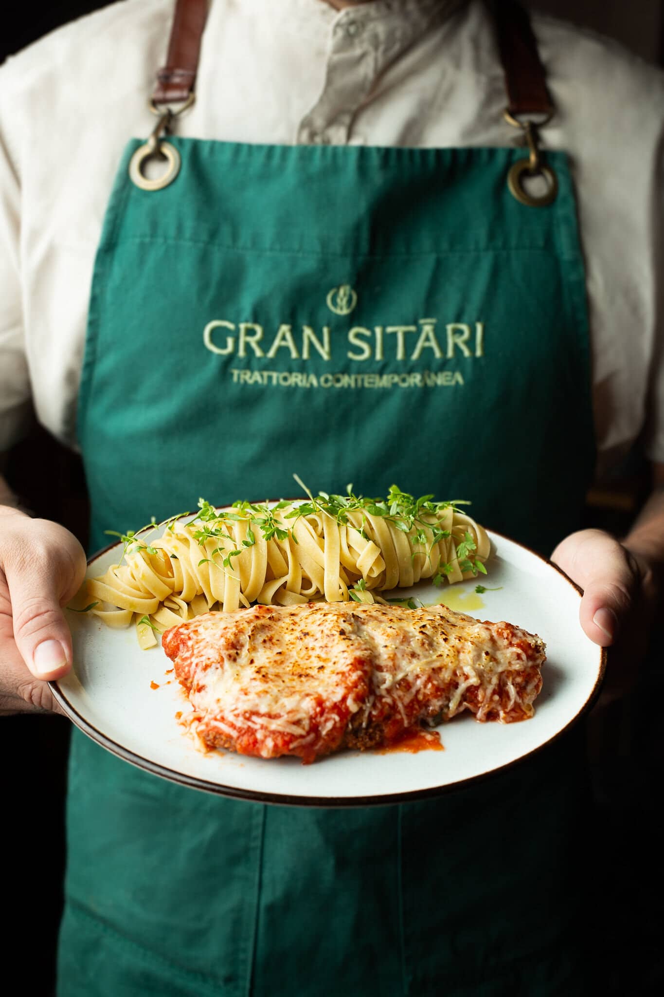 Cozinheiro de avental verde do Gran Sitári segurando um prato de parmegiana com tagliatelle.
