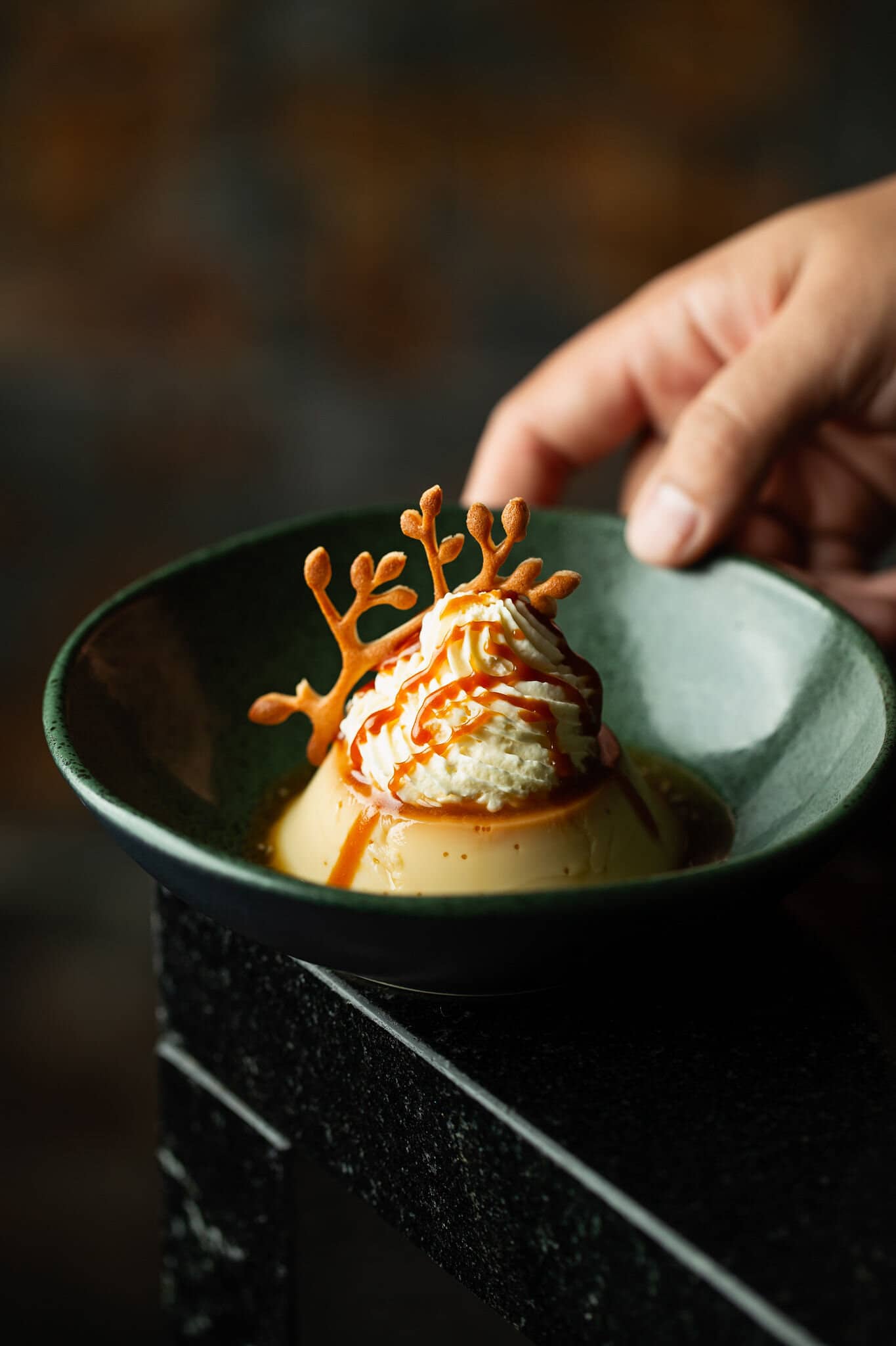 Sobremesa com creme e calda de caramelo e uma tuile dourada, em prato verde escuro.