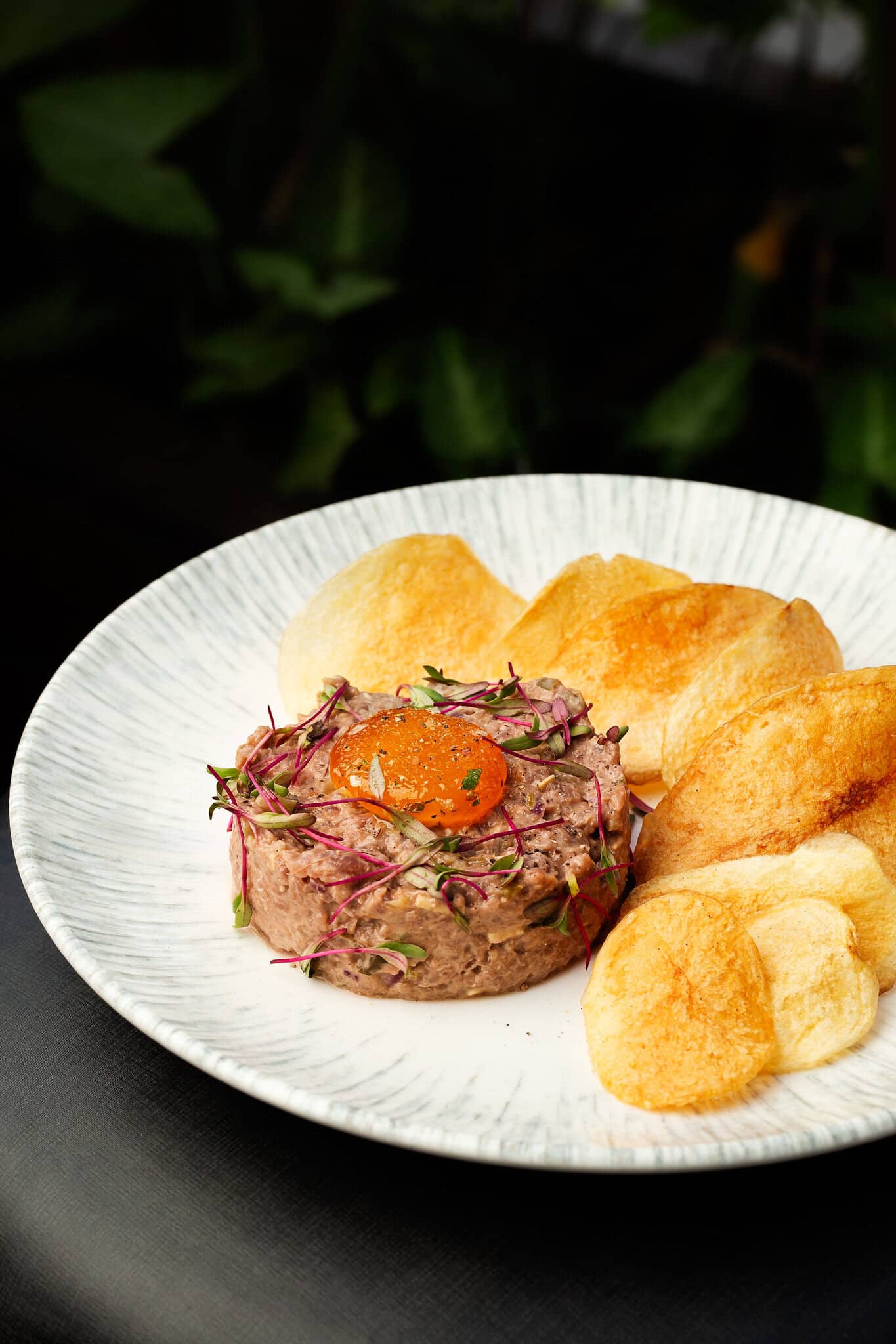 Tartar de filé com gema curada e chips de batata em prato branco riscado.